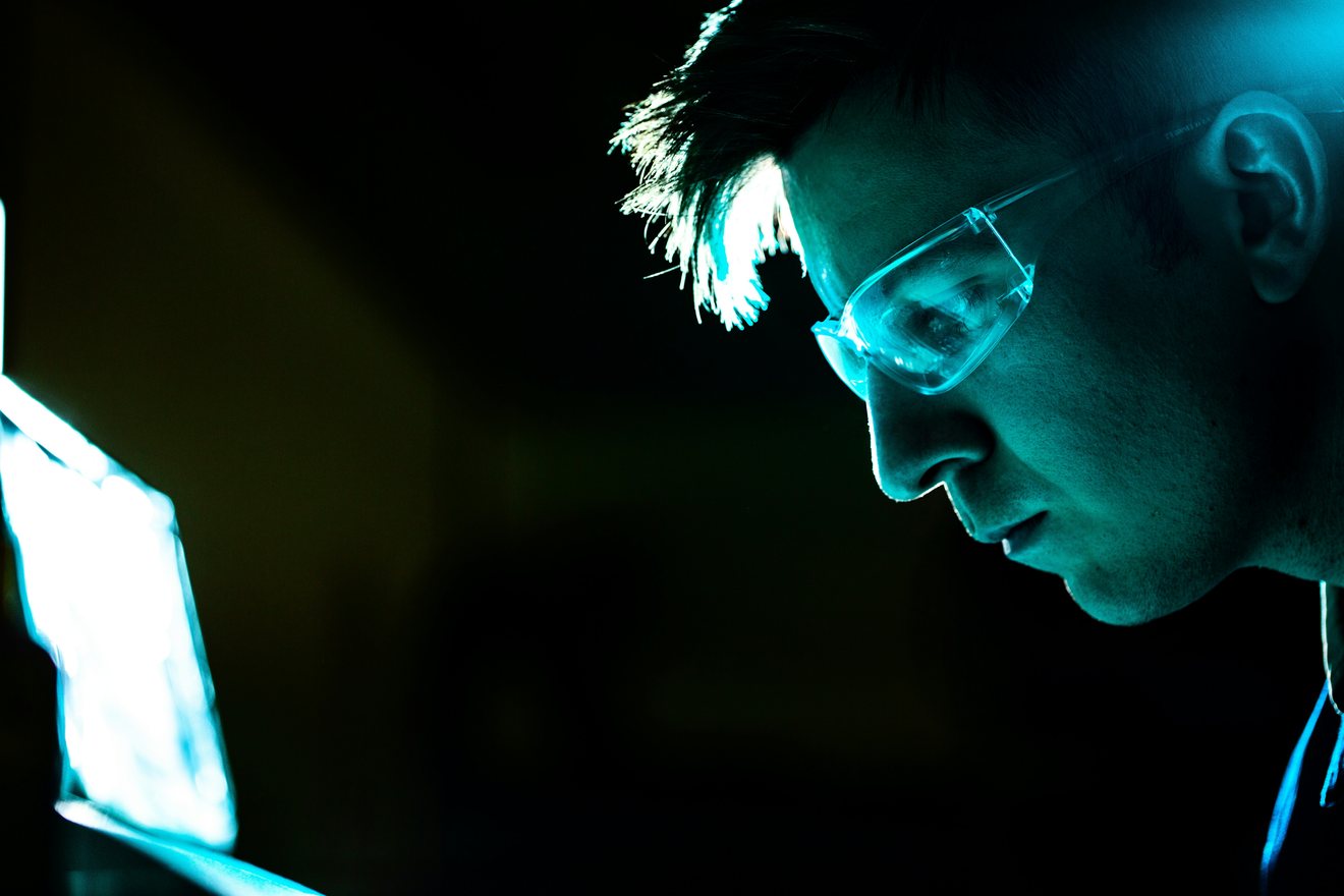 An engineer in safety glasses studying an illuminated screen in a darkened workspace