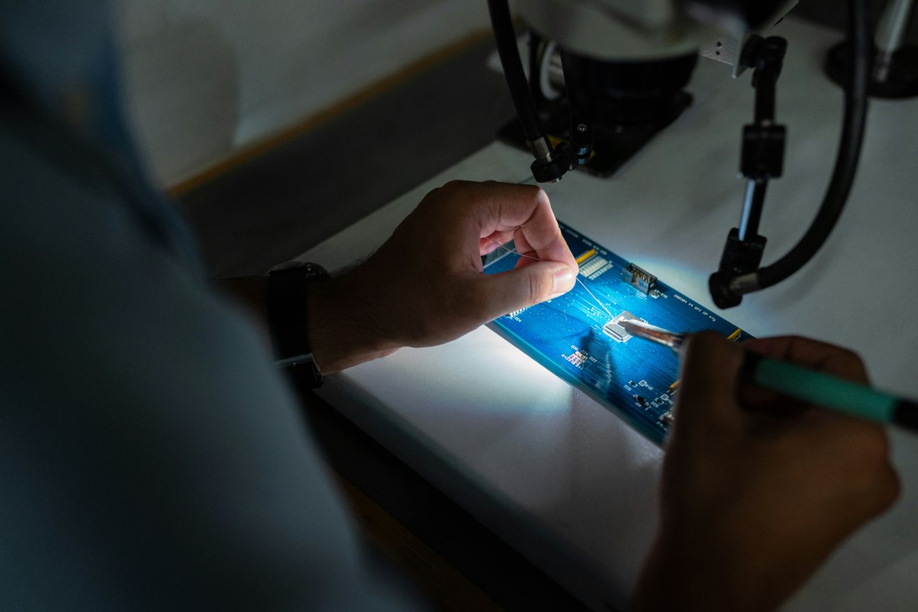 Close-up of hands fitting a fine wire to an illuminated circuit board under a bench microscope