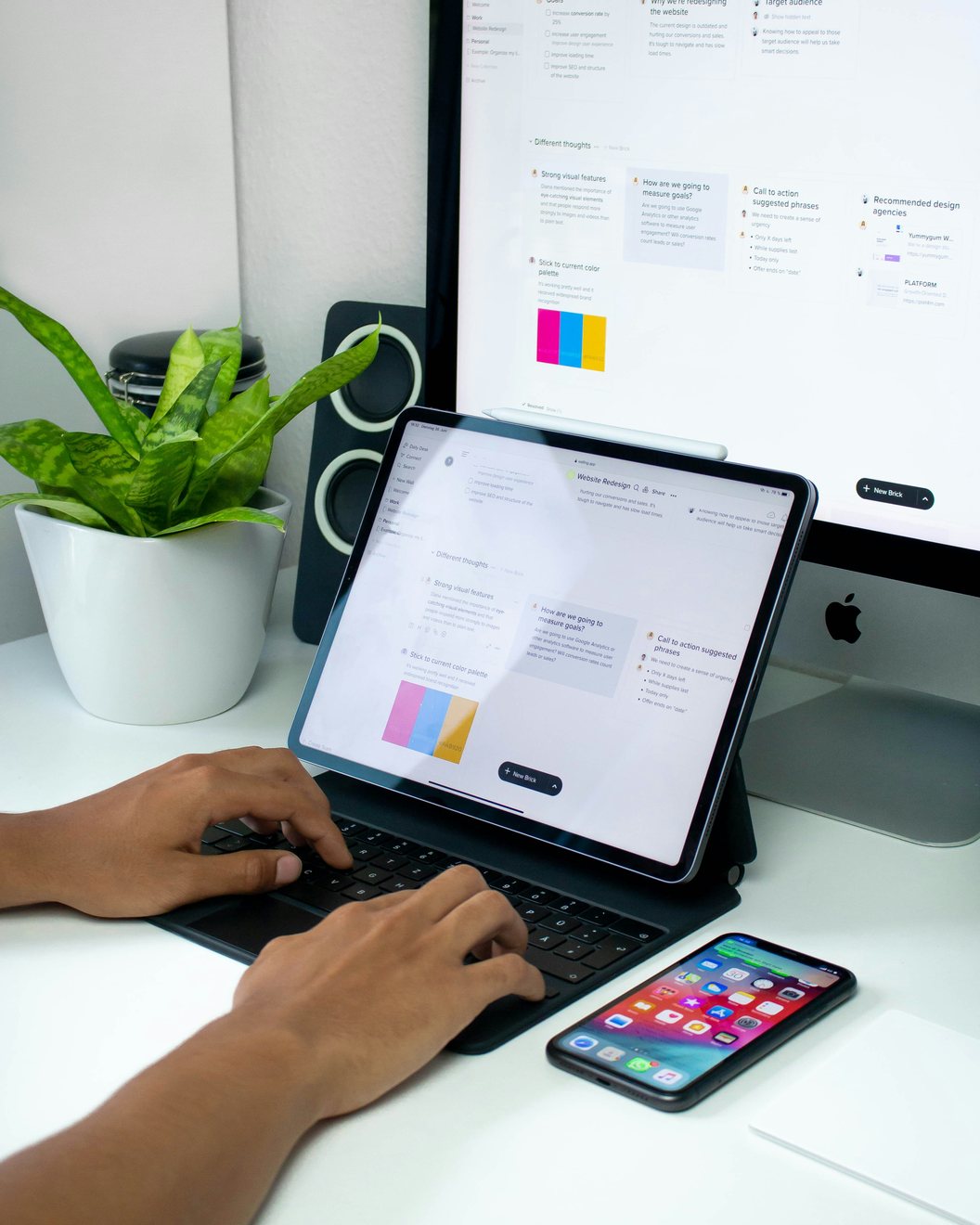 Hands typing on a tablet keyboard at a white desk with a project board displayed on a larger monitor behind