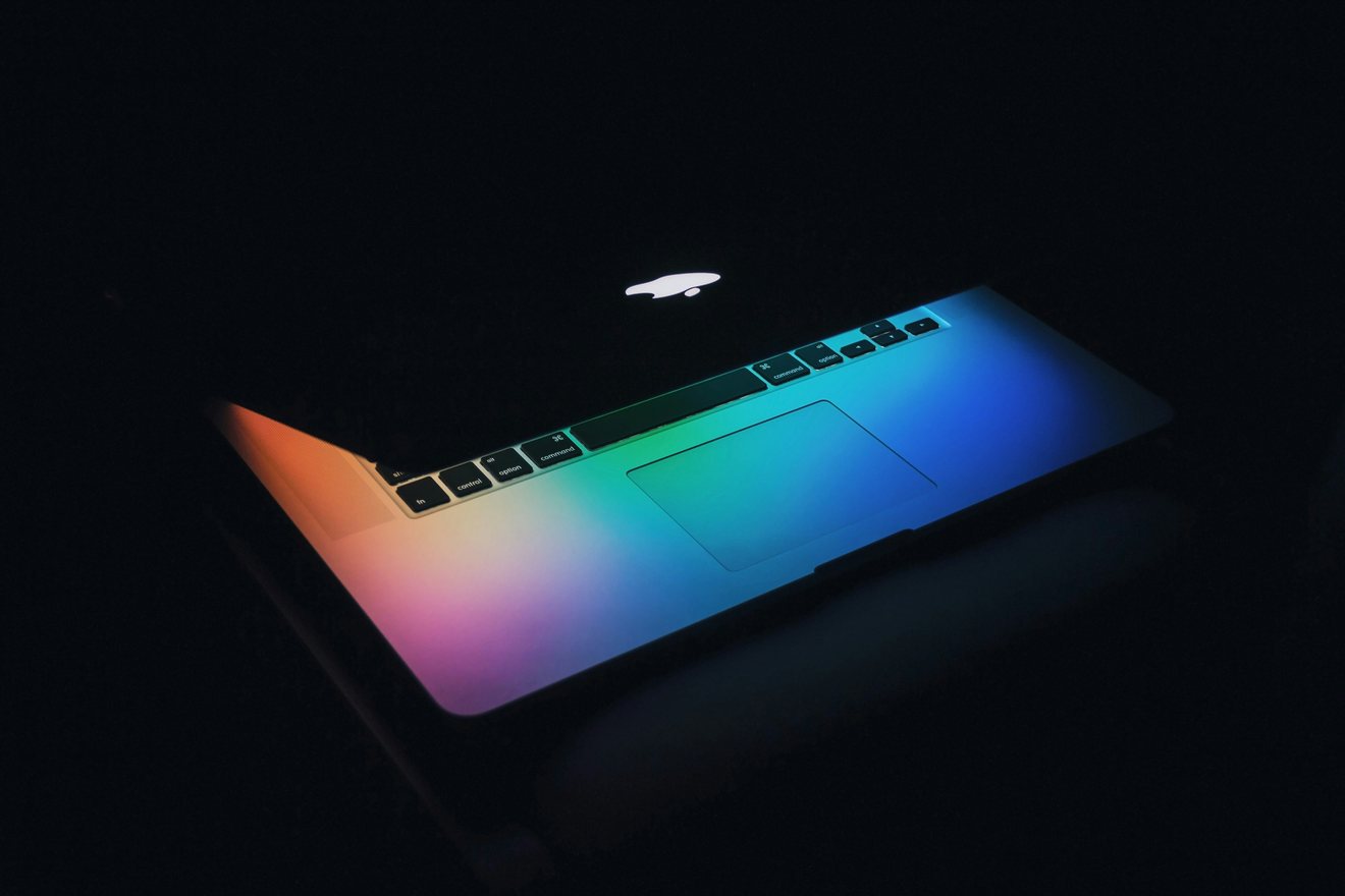 An open laptop in a darkened room lit by a rainbow gradient spilling across its keyboard and trackpad